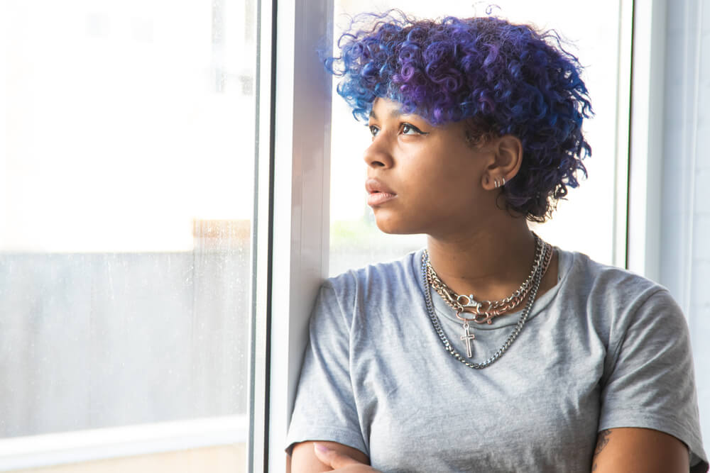 curly blue afro young woman looking out window in contemplation about not feeling happy yet not feeling sad