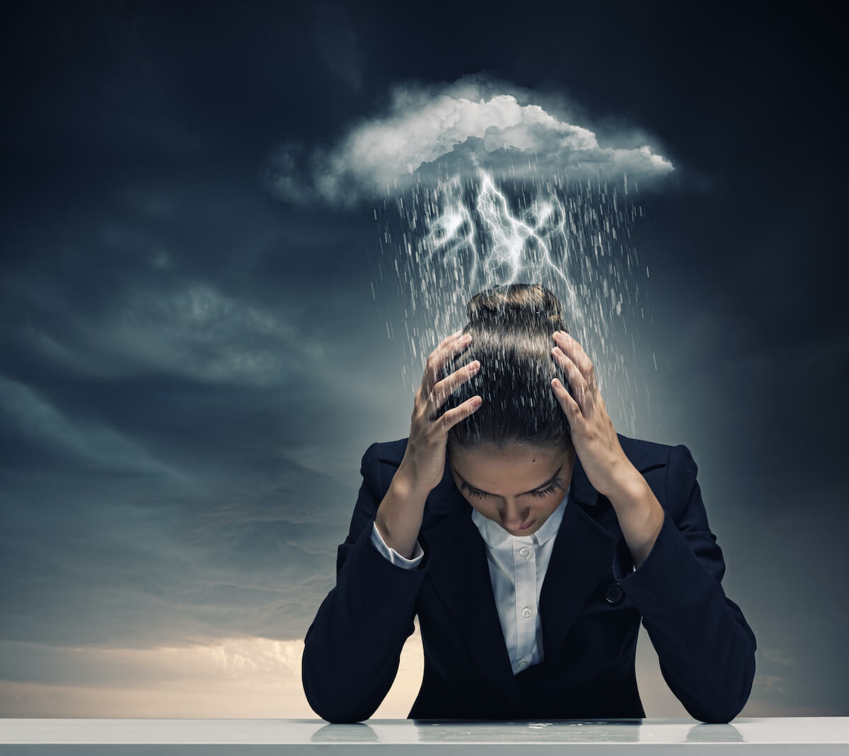 anxiety ridden business woman leaning over a desk with hands in her head and a stormy cloud above seeking relief from her anxiety
