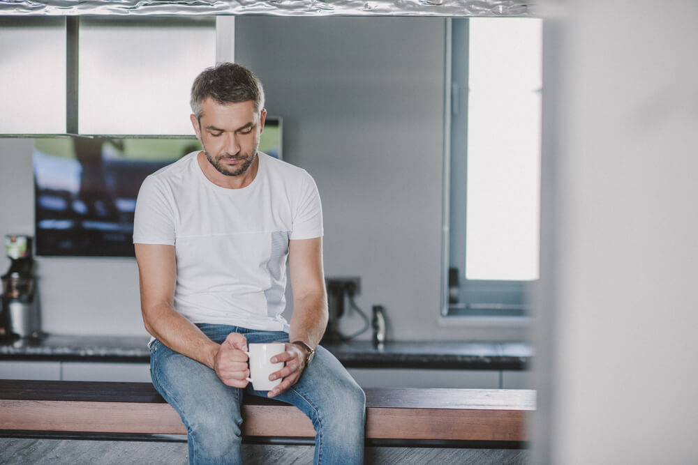 white guy sitting on counter holding cup looking downwards wondering why if he doesn't feel sad why he's not happy